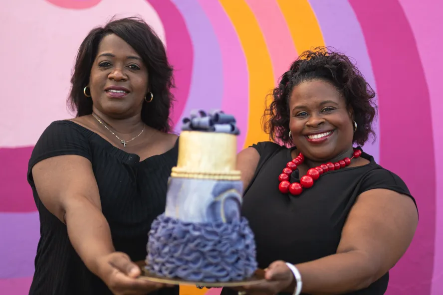 Two women hold up an elaborately decorate cake in front of a bright pink mural
