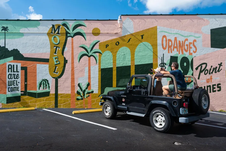 A man and woman in a black jeep park in front of a vintage-themed mural with signs for "motel," "oranges" and "all are welcome here"