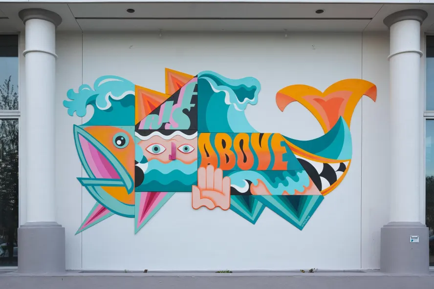 A bright blue, orange and pink mural of a fish created in geometric designs with shapes of a hand and face inside says Rise Above