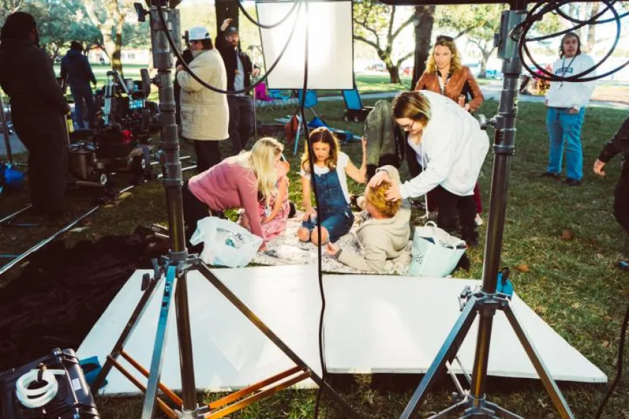 A family sits on a blanket on the grass outside while people setup cameras, hair, and makeup.