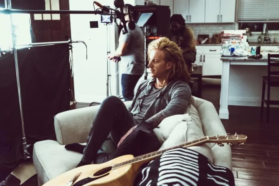 The singer sits on a couch next to a guitar with a filming crew in the background.