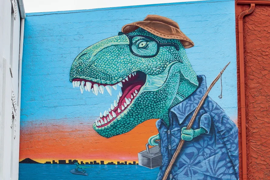 a mural depicts a tyrannosaurus rex dressed as if he is going fishing with a fishing pole, hat and tackle box. In the water behind him, a smaller dinosaur rides a jet ski