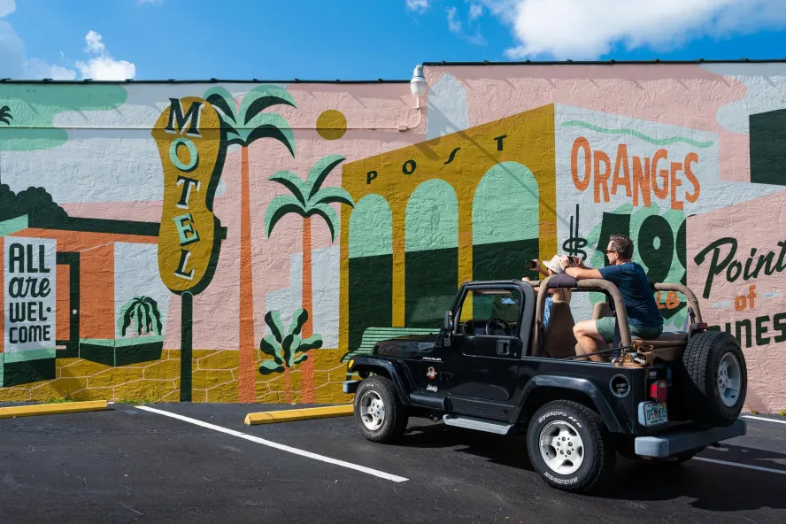 A man and woman in a black jeep park in front of a vintage-themed mural with signs for "motel," "oranges" and "all are welcome here"