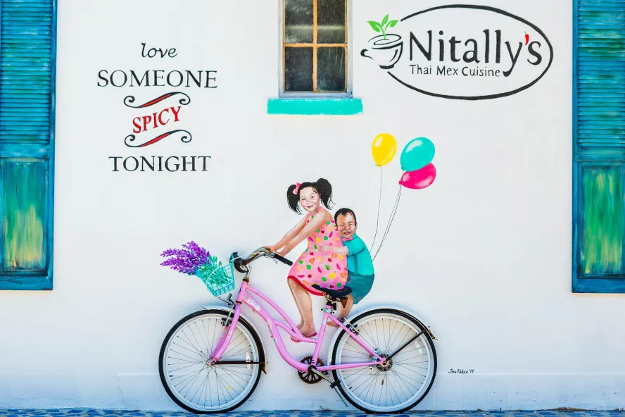 Colorful painting on a white wall of two kids riding a pink bike with balloons on the back