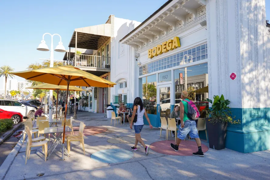 Two people in casual summer clothes walk past bodega in the EDGE District of St. Pete. Tables with yellow umbrellas are on the sidewalk