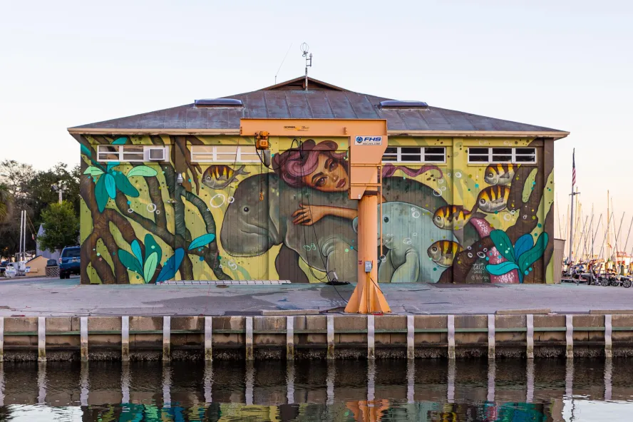 a mural on the side of a building depicts a woman with swirling reddish hair riding or hugging a manatee while her calf and fish swim nearby