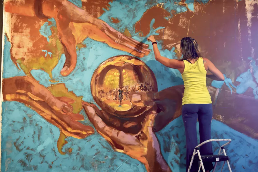 artist Carrie Jadus in a yellow shirt and bluejeans standing on a ladder painting a mural in st. pete