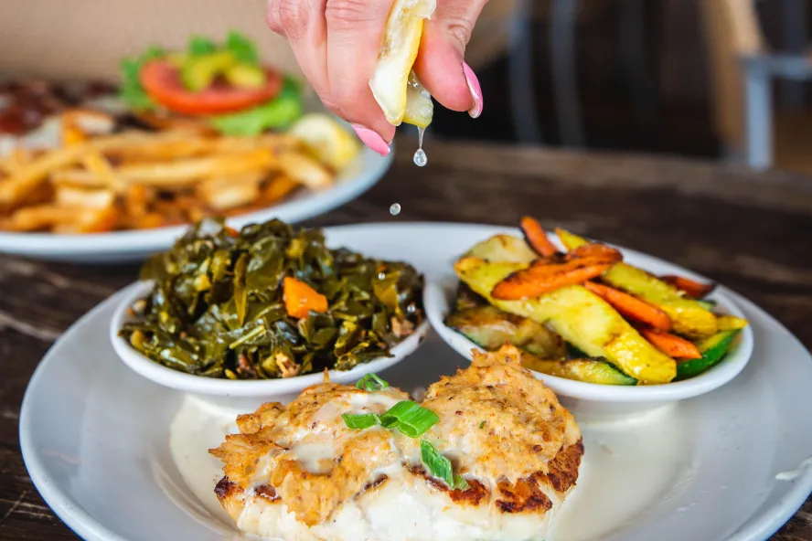 a woman squeezing a lemon on top of a plate of grilled fish, sauteed greens and mixed veg at Hog Island Fish Camp in Dunedin