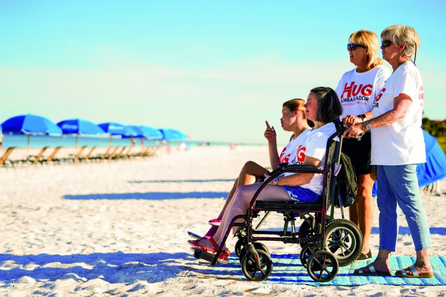two women in white t-shirts and jeans accommodate two young girls in wheelchairs on a beach access mat in Indian Rock Beach, blue umbrellas are in front of them on the sand