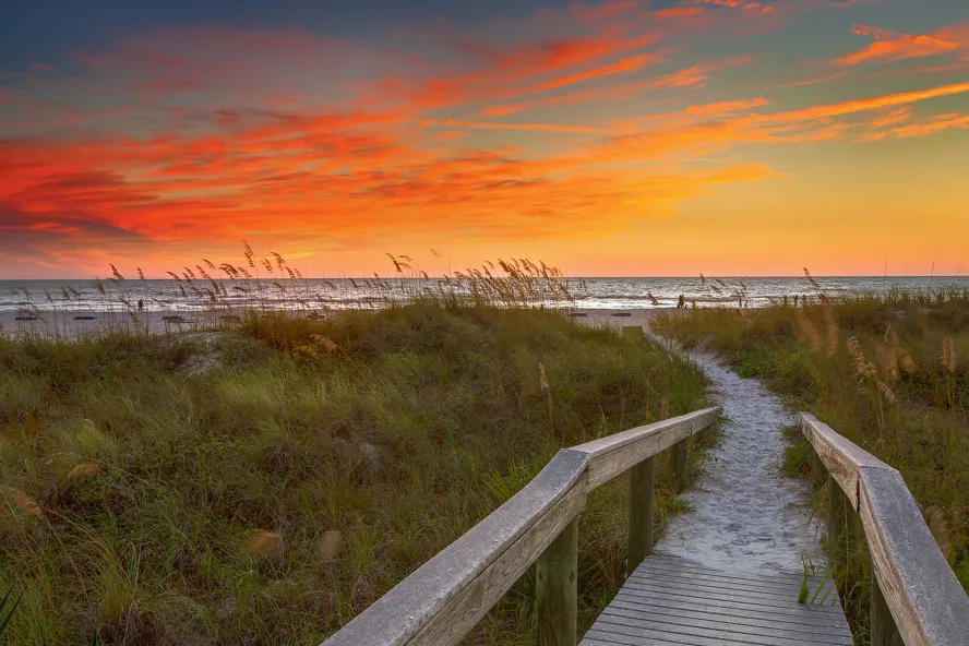 a wooden boardwalk through sea oats and other vegetation leads to the Gulf, the boardwalk is sandy, the sky is just after sunset with orange and blue colors