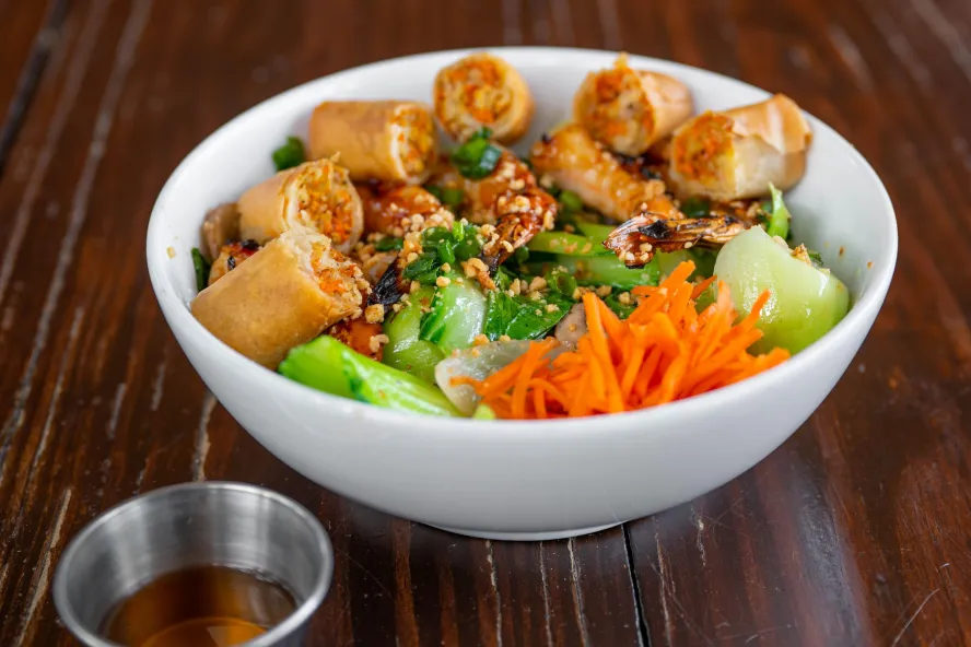 a large white bowl filled with egg rolls, peanut crumbles and bok choy on a wooden table at Alesia in St. Pete