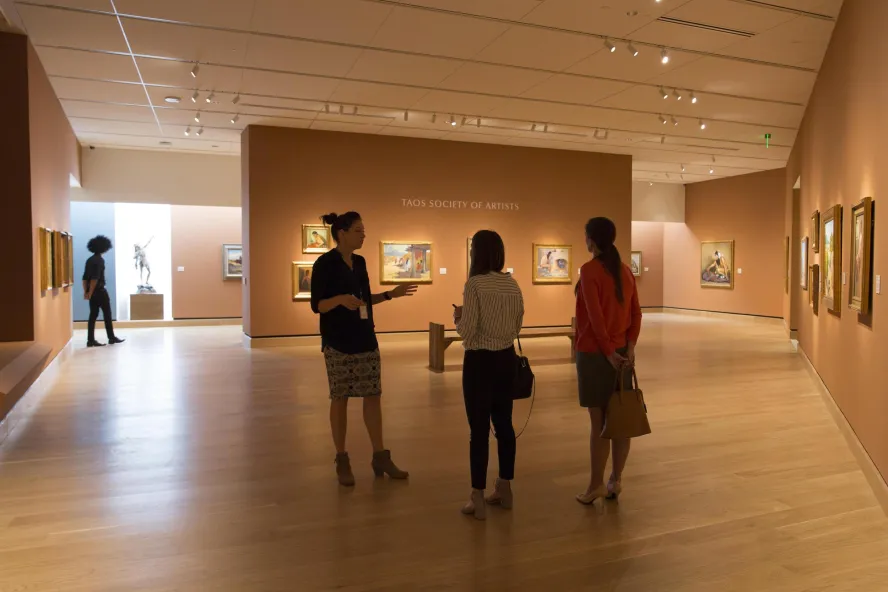 Three people talk in a gallery of paintings at the James Museum.