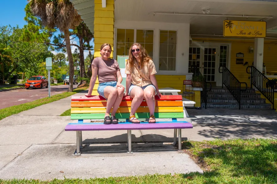 two women in shorts and tshirts sit on top of a bench painted in rainbow colors in front of a bright yellow inn next to a street
