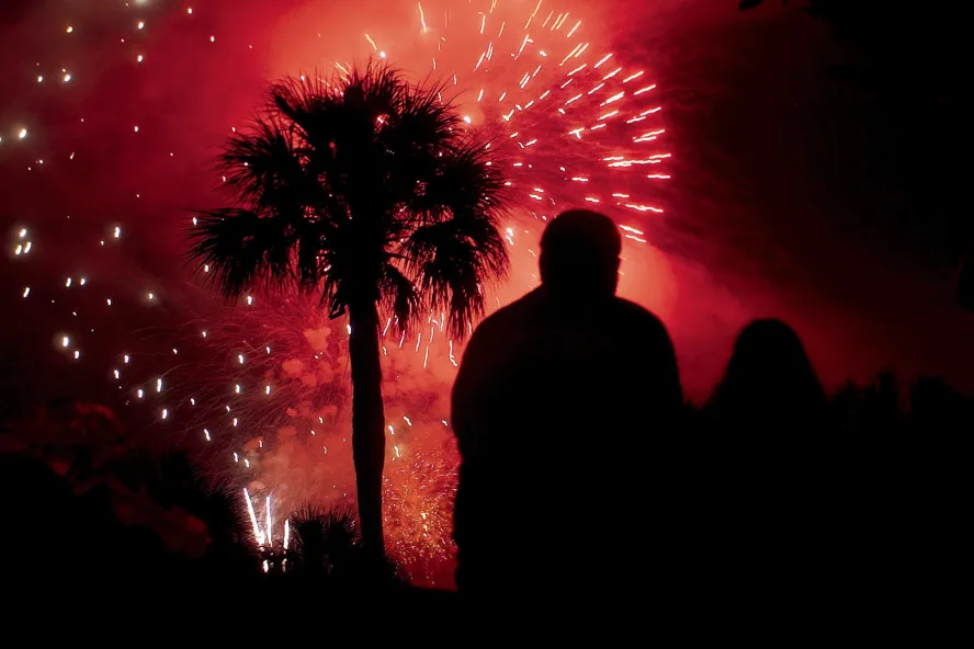 Fogos de artifício explodem no céu vermelho com silhueta de palmeira e duas pessoas