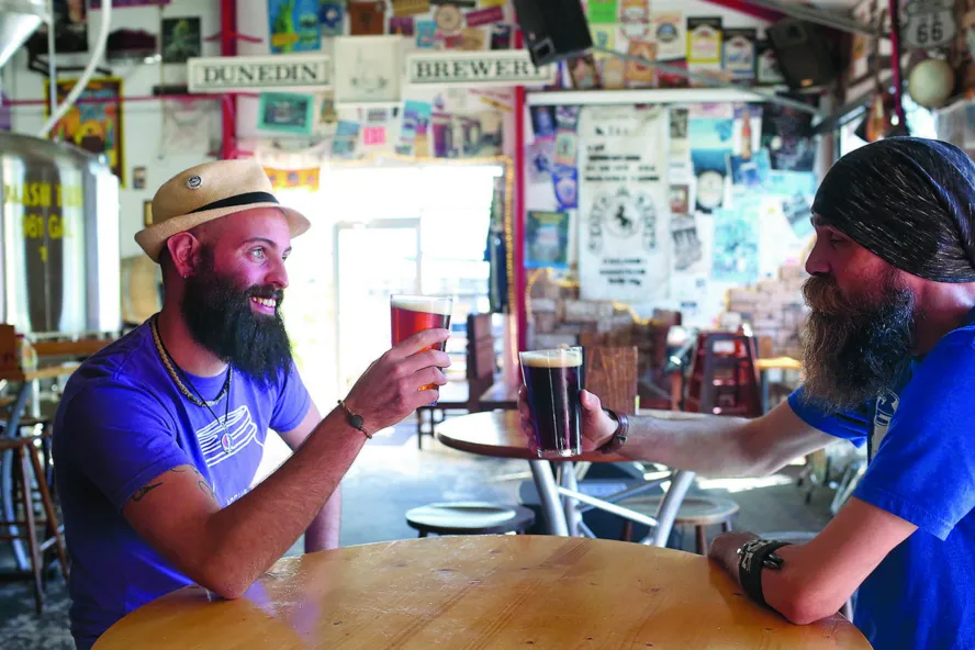 Two men in blue shirts toast with beer. Walls are covered with stickers.