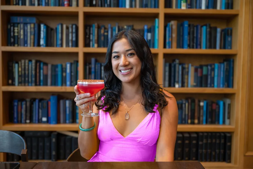 a woman in a Barbie pink dress smiles and holds up a pink cocktail with a background of bookshelves at The Library restaurant