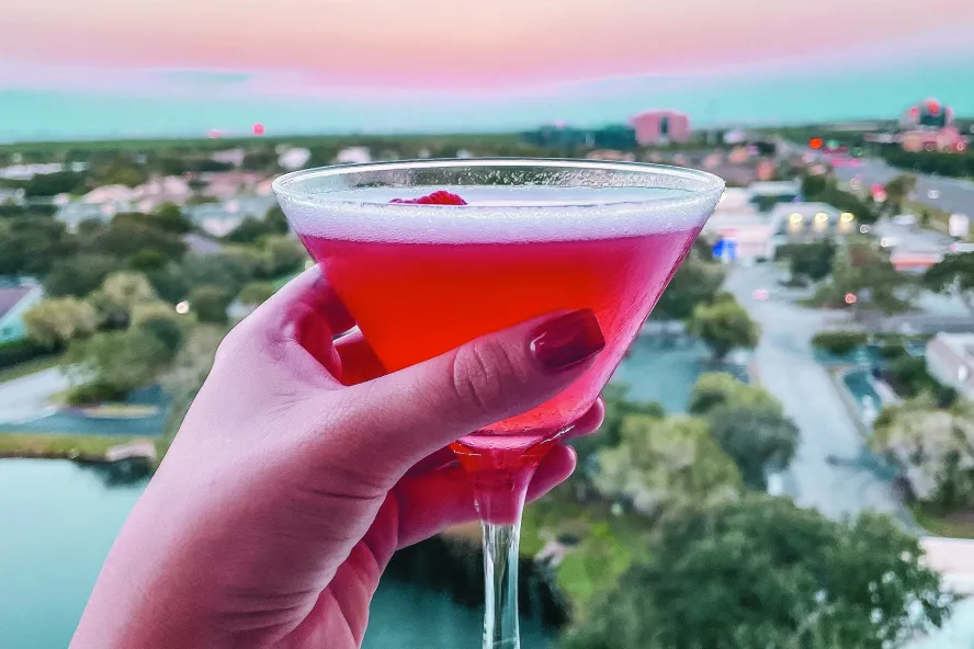 a manicured hand holding a pink cocktail with a cherry with a view of sunset and the Gulf from Vantage Rooftop Bar at the Karol Hotel