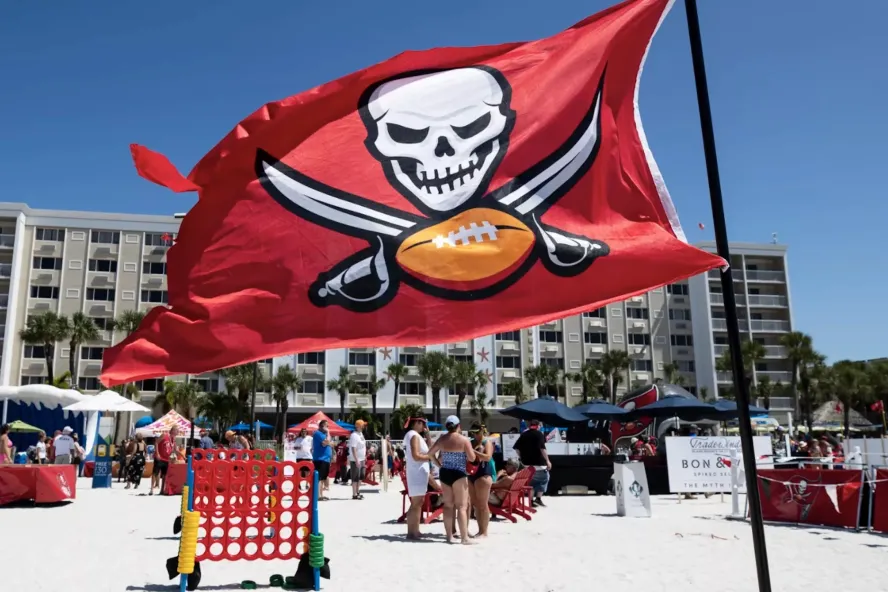 A group of people standing on a beach playing games and mingling with the Tampa Bay Bucs red flag blowing in the wind.