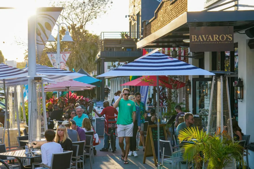 Pedestrians walk through sidewalk restaurant tables and umbrellas