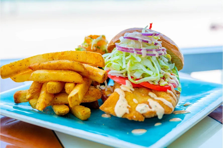 fried grouper sandwich with onions lettuce tomatoes and french fries on a bright blue plate