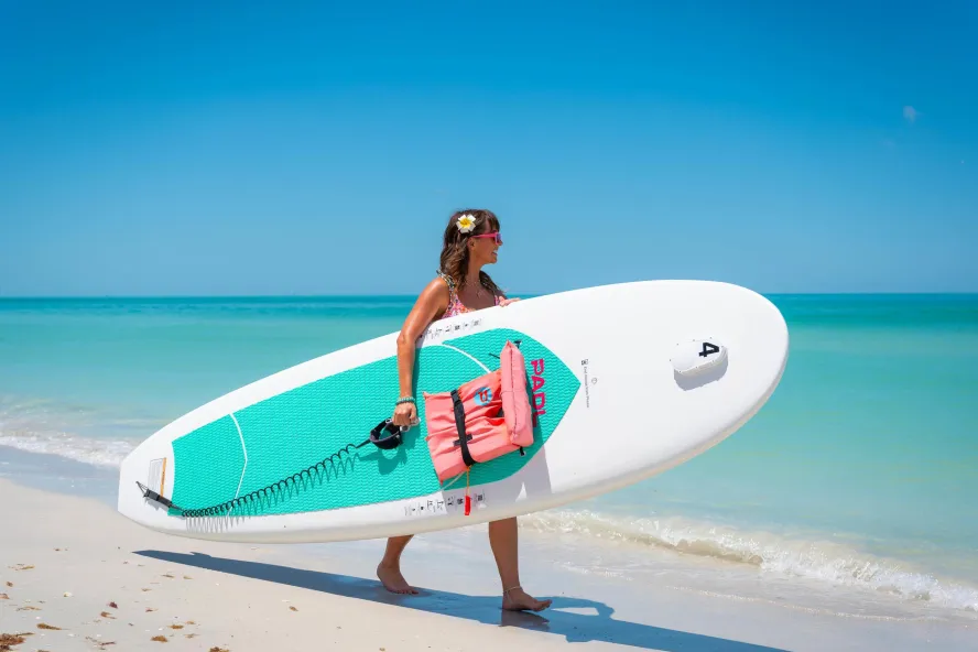 Woman walks along the ocean holding a stand-up paddleboard under her arm.