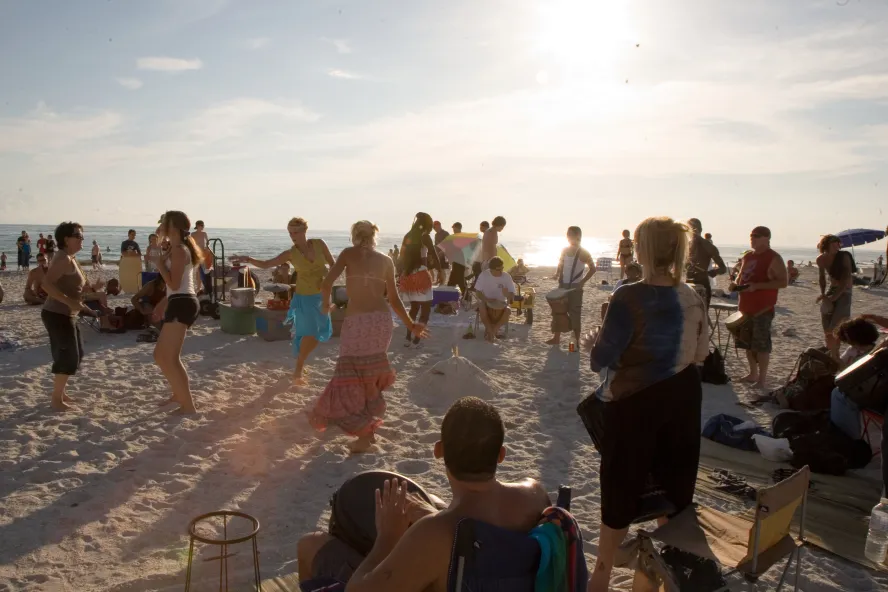 A group of beach-goers dance in the sand to drums on the beach at sunset.