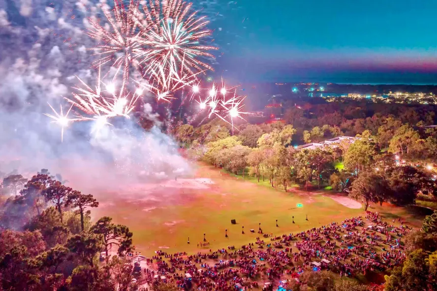 Independence Day fireworks in a park with a crowd of people watching during the evening.