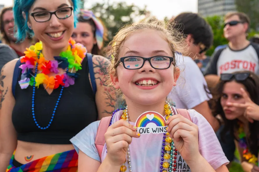 a smiling girl wearing glasses and rainbow beads holds a "love wins" rainbow sign while standing next to her mom