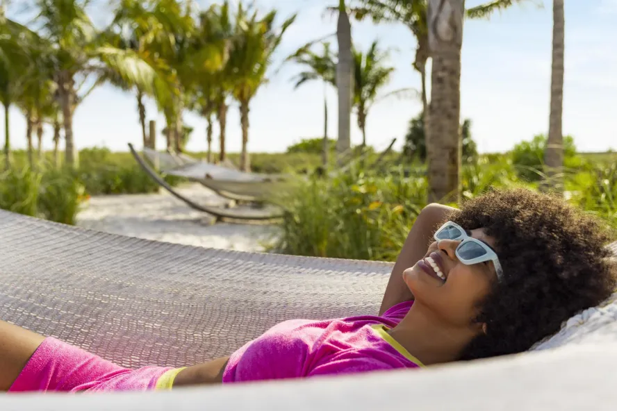 Woman with sunglasses laying in a hammock with the beach and palm trees in the background