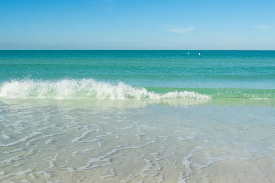 a small wave rolls up on a white beach, the Gulf water is turquoise and clear and the sky is blue