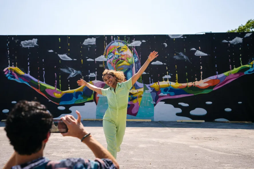 a person taking a photo of a woman wearing a lime green jumpsuit with her arms outstretched in front of a mural of a black woman with her arms outstretched