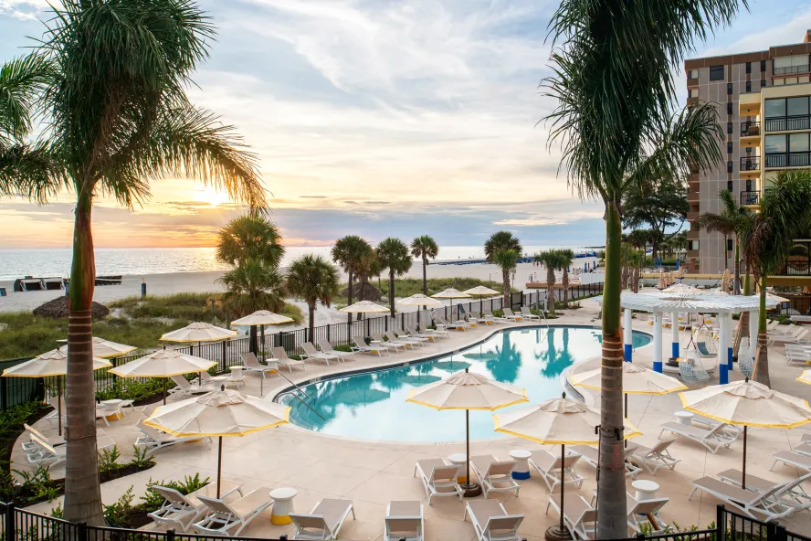 palm trees in foreground and a pool with chairs and umbrellas around it near the beach at sunset at Sirata Resort