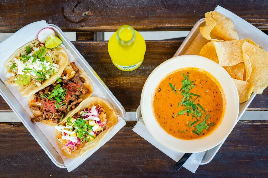 a tray with three kinds of tacos next to a tray with mexican soup and chips, with a yellow soda in the middle