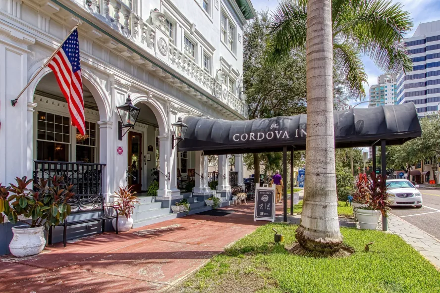 the white facade of the Cordova Inn with a huge black awning and an American flag, next to large palm trees, brick walkway and grass