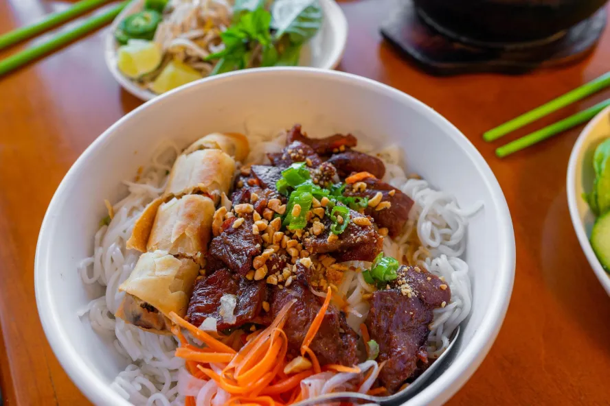 a bowl with white rice, a spring roll, an asian beef entree and decorated with pickled vegetables