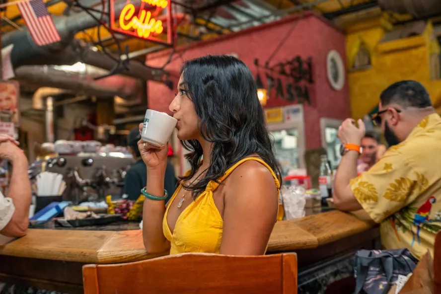 a young woman in a yellow sundress sips coffee at a wooden counter with other patrons at a restaurant