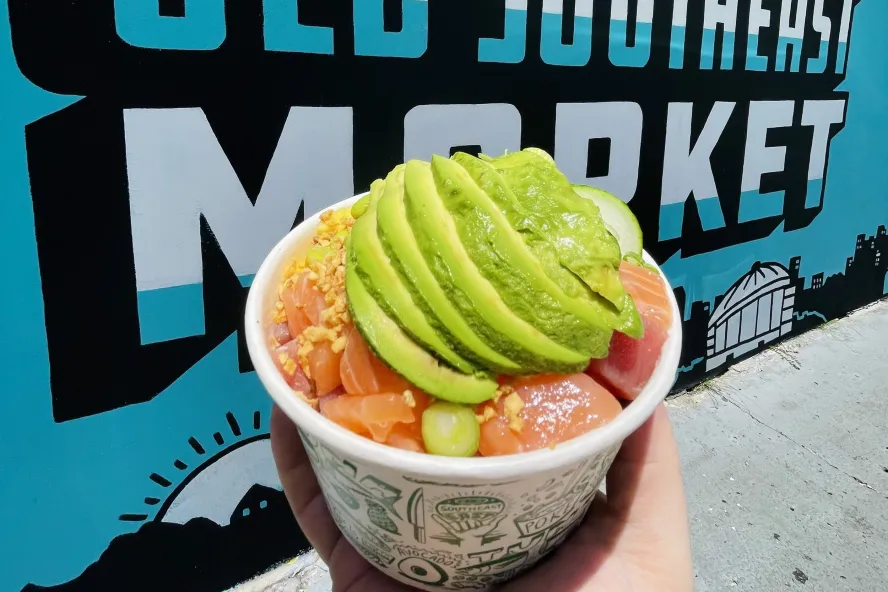 a hand holding a paper bowl with poke topeed with guacamole swirl