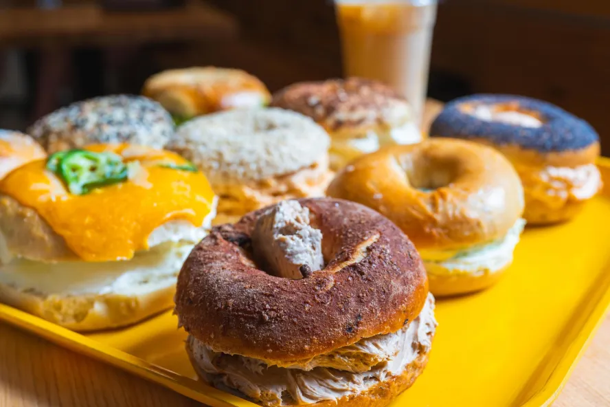 a yellow tray holding bagel sandwiches of many kinds, with an iced coffee in the background