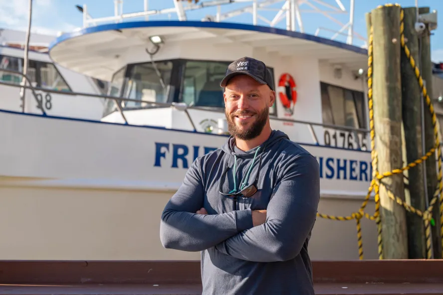 a smiling man with a beard wearing a baseball cap standing in front of a large fishing boat