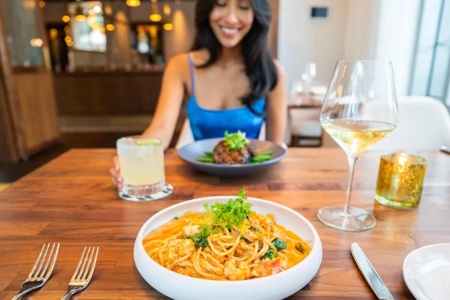 a smiling woman in a bright blue dress sits across from a bowl of pasta at a restaurant