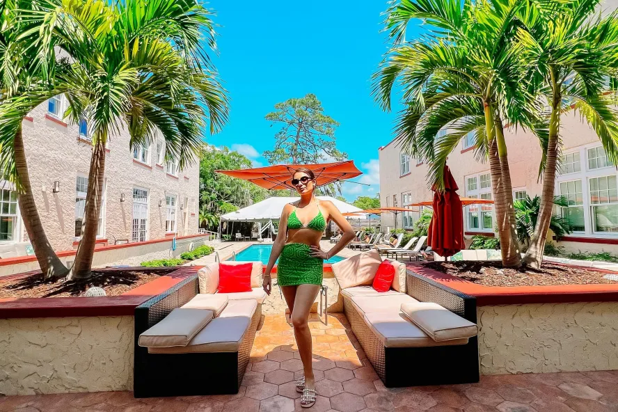 a woman in a bright green bathing suit posing in front of benches and a pool near palm trees at the Fenway Hotel