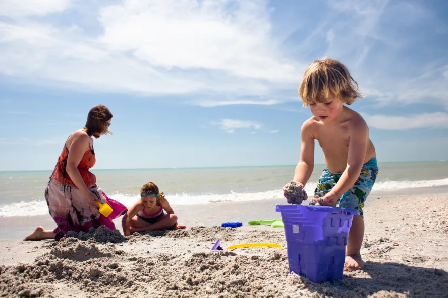 a little boy puts sand in a large purple bucket shaped like a sandcastle while his mom and sister play in the sand behind him on the beach