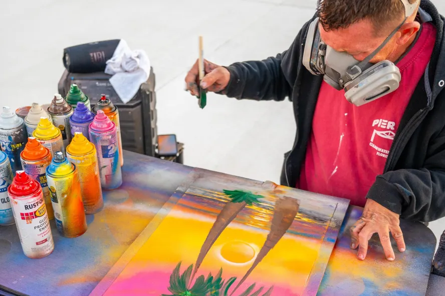 an artist wearing a heavy-duty mask uses airbrushed paints to create a palm tree painting at the Sunsets at Pier 60 celebration