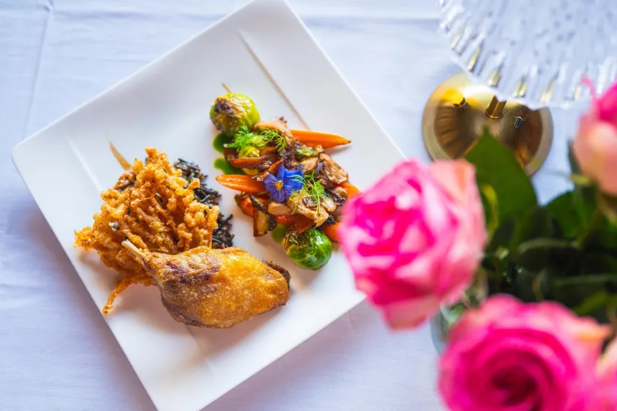 A view of a beautifully plated dish at Black Pearl Restaurant in Dunedin, accompanied by bright pink roses on the white linen tablecloth