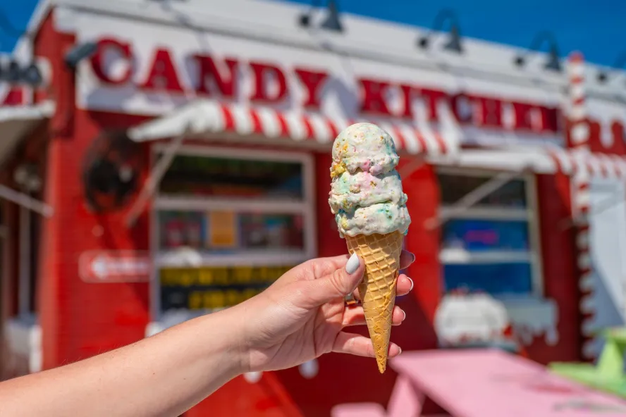 a person's hand holding a stacked ice cream cone in front of the nostalgic red and white Candy Kitchen in Madeira Beach