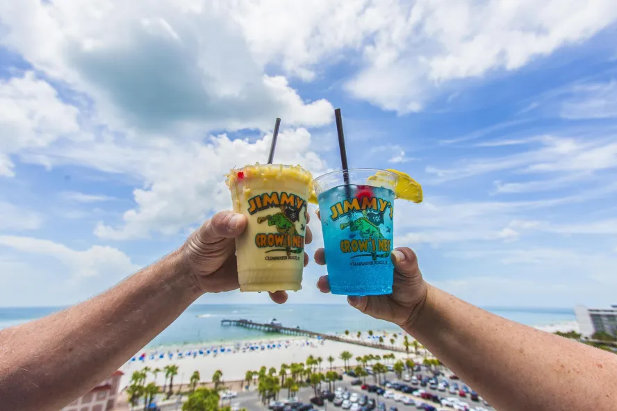 two tropical drink in a Jimmys Crow's Nest plastic cup cheersing with rooftop view in background of Clearwater Beach and Pier