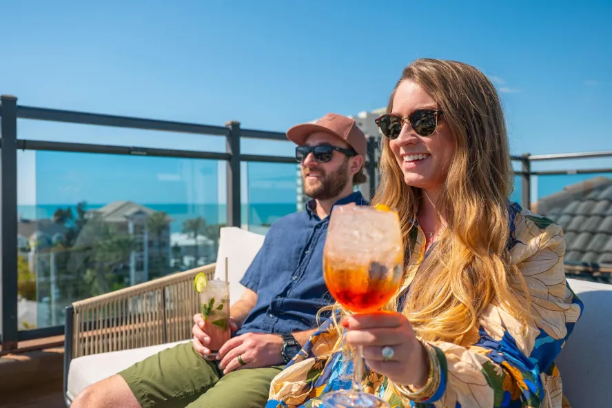 woman and man holding cocktails sitting outside on rooftop smiling