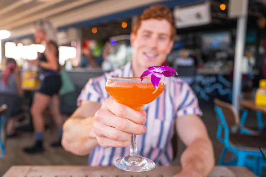 man holding colorful cocktail up with flower on rim at an outdoor restaurant
