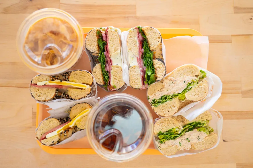 a light wood table with a tray of bagel sandwiches and iced coffee drinks on top, shot from above