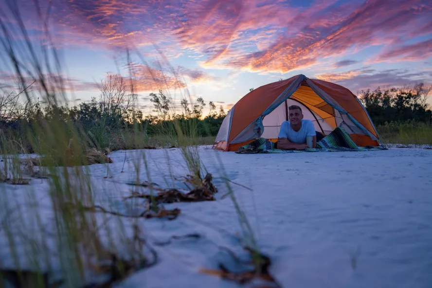 a young man relaxes inside his tent on the beach at Shell key with a dramatic pink and blue sky behind him